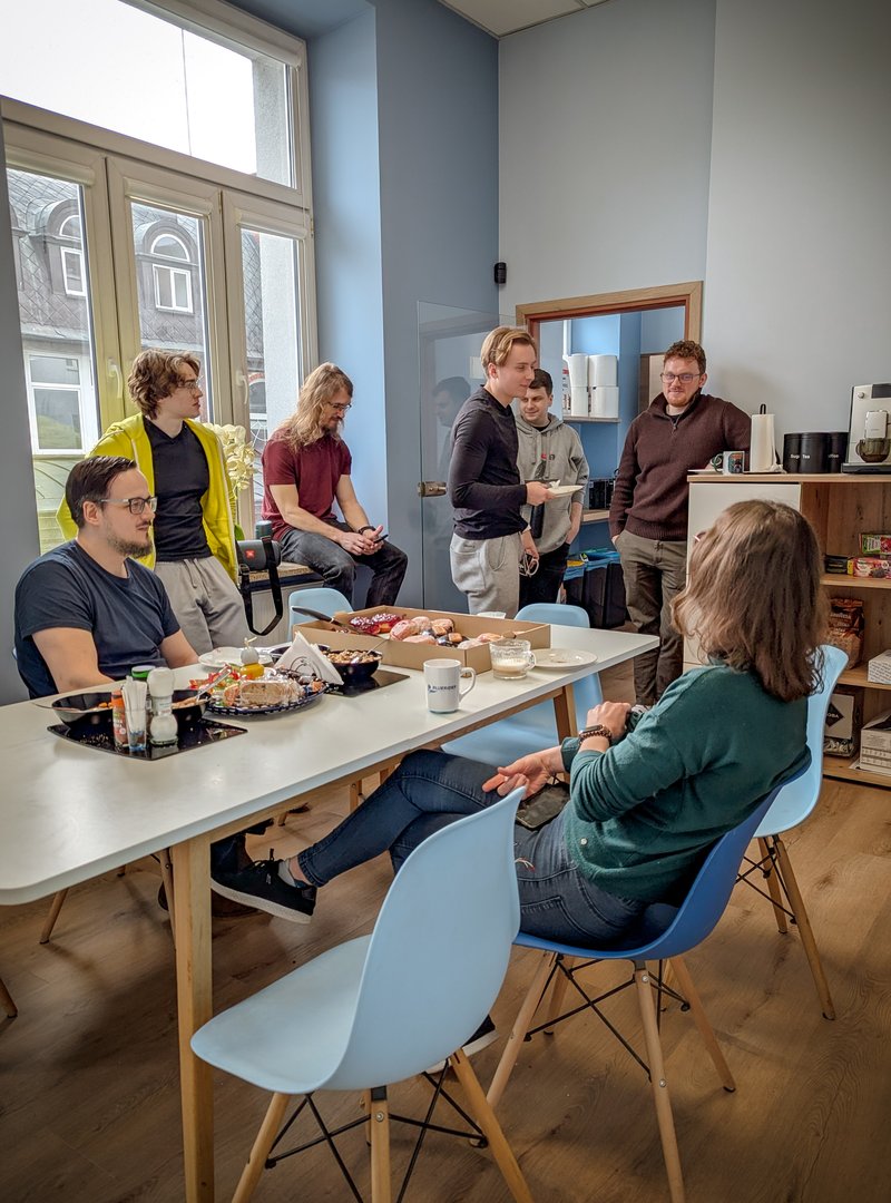 Grupa osób prowadzi ożywioną dyskusję w kuchni biurowej w coworkingu BlueRider.Software. Na stole przed nimi stoją kubki z kawą i jedzenie, a atmosfera spotkania jest przyjazna i nieformalna.