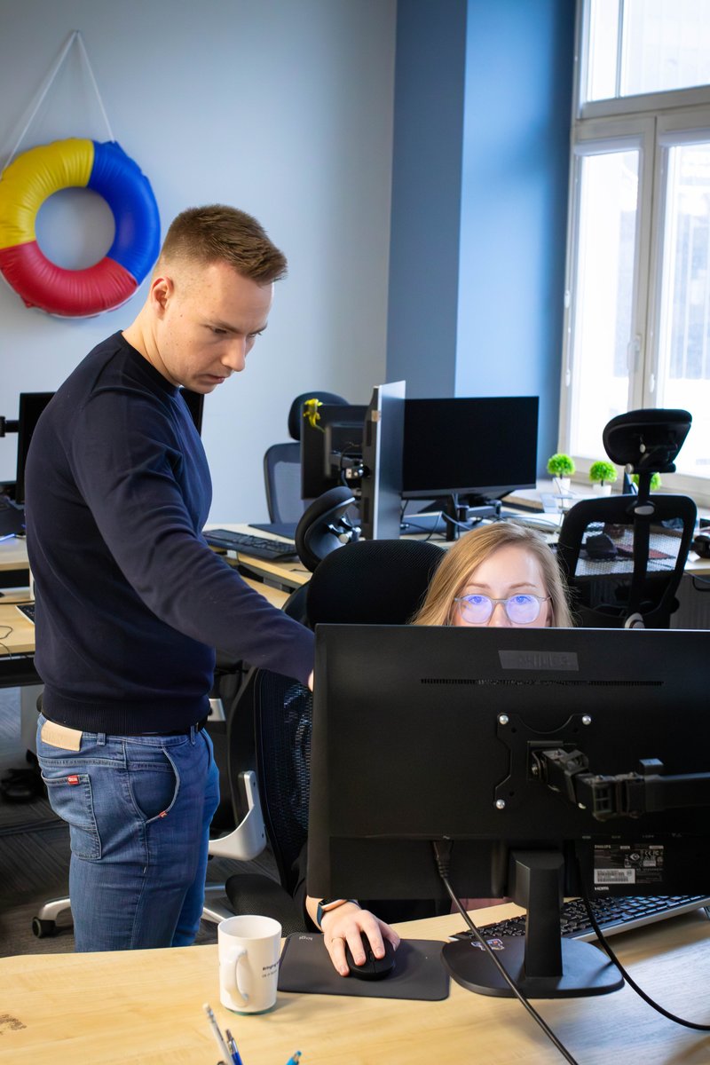 Dwóch programistów z zespołu BlueRider.Software współpracuje przy biurku w biurze. Mężczyzna stoi i wskazuje coś na monitorze komputera kobiecie, która siedzi przy biurku.