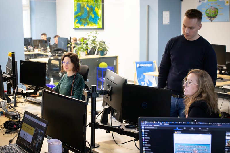 Mentor w biurze BlueRider.Software pomaga dwóm koleżankom przy pracy. Mężczyzna stoi za nimi i patrzy na monitor komputera, a kobiety siedzą przy biurkach w otwartej przestrzeni biurowej.
